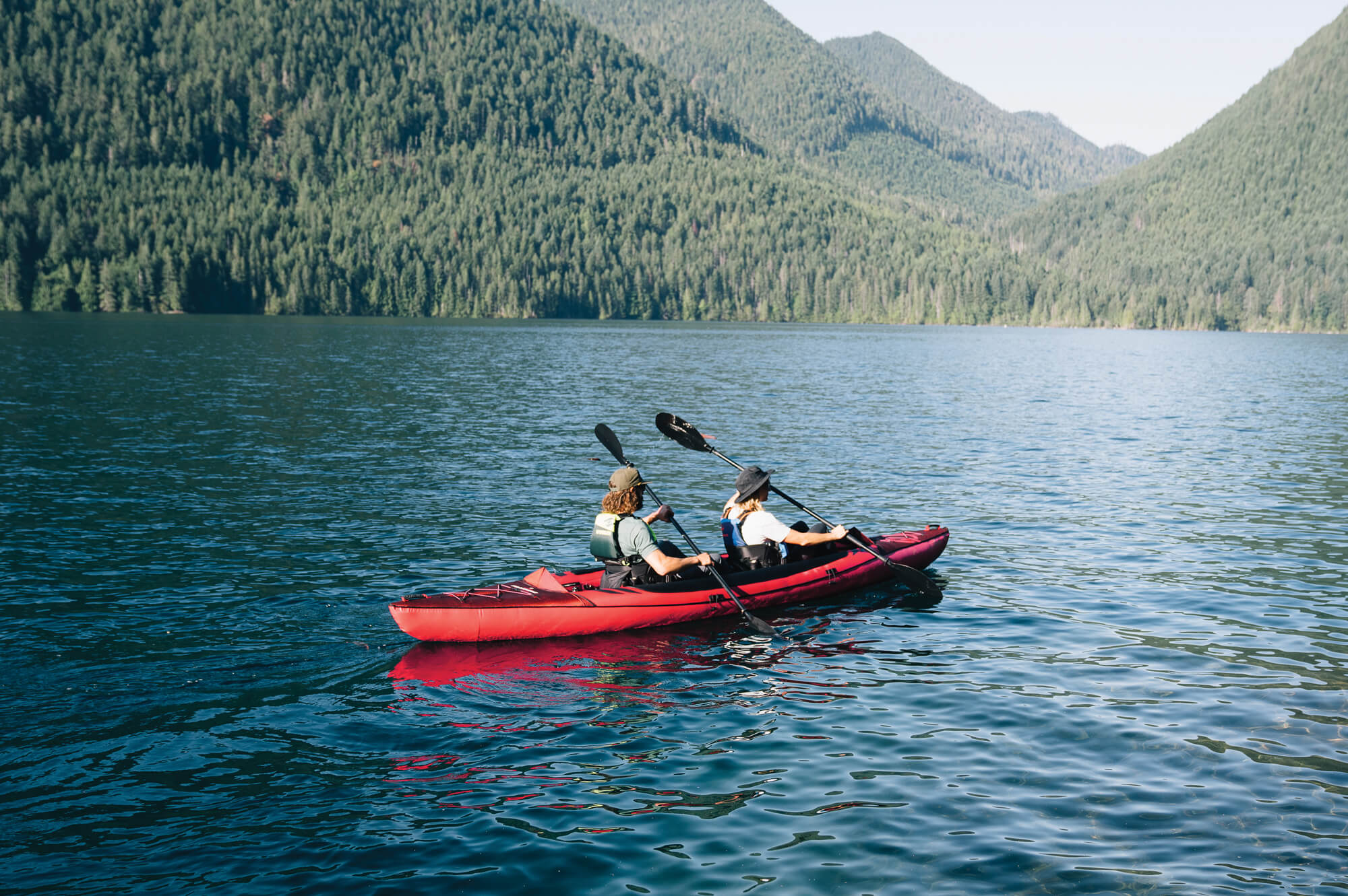 Two people paddling across a mountainous lake on a red, two-person kayak from HO Sports.