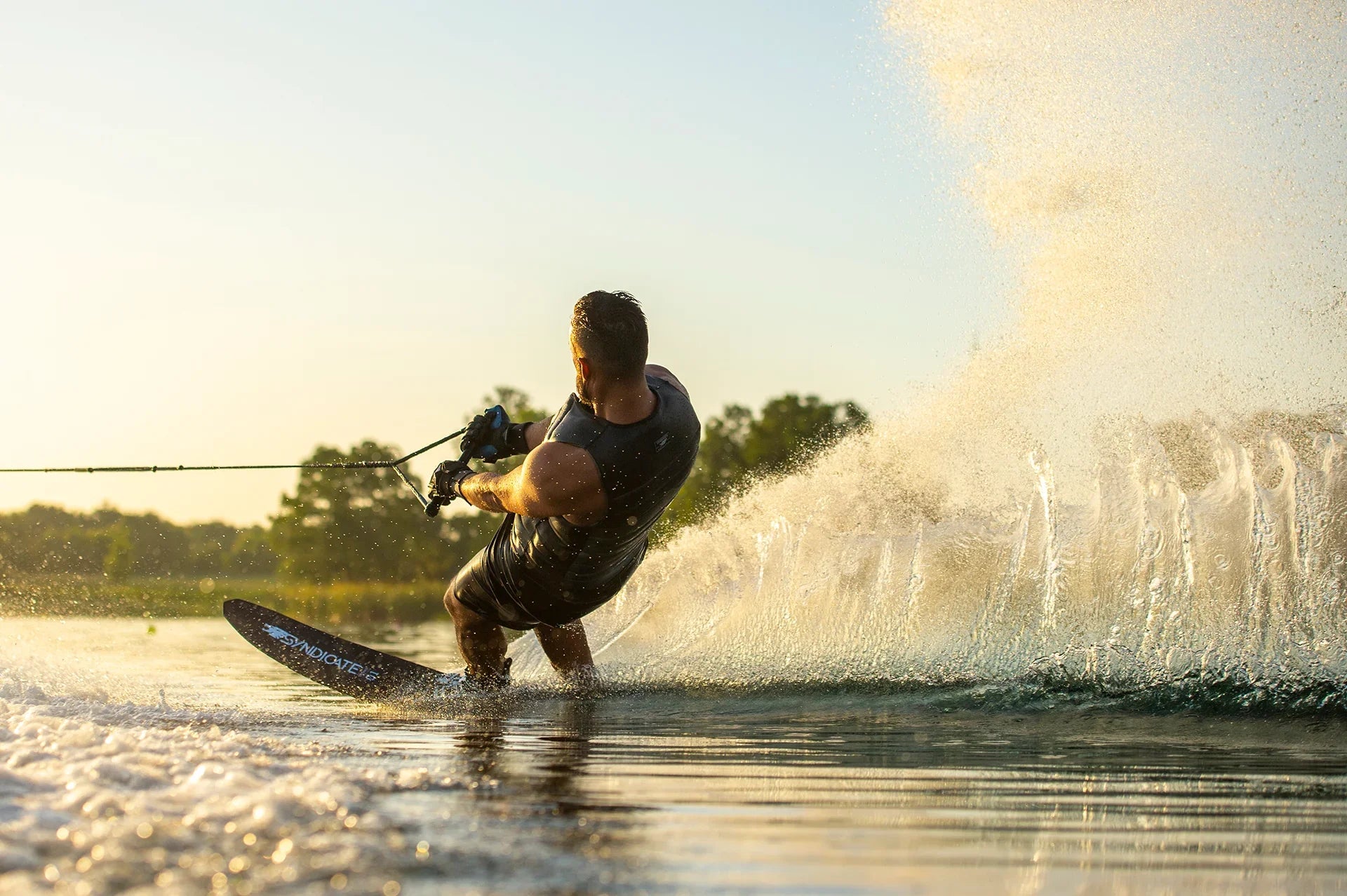 Water Skier leaning back to turn splashing up water at sunset wearing a black HO Sports life vest and riding a black Syndicate Ski with a white logo.