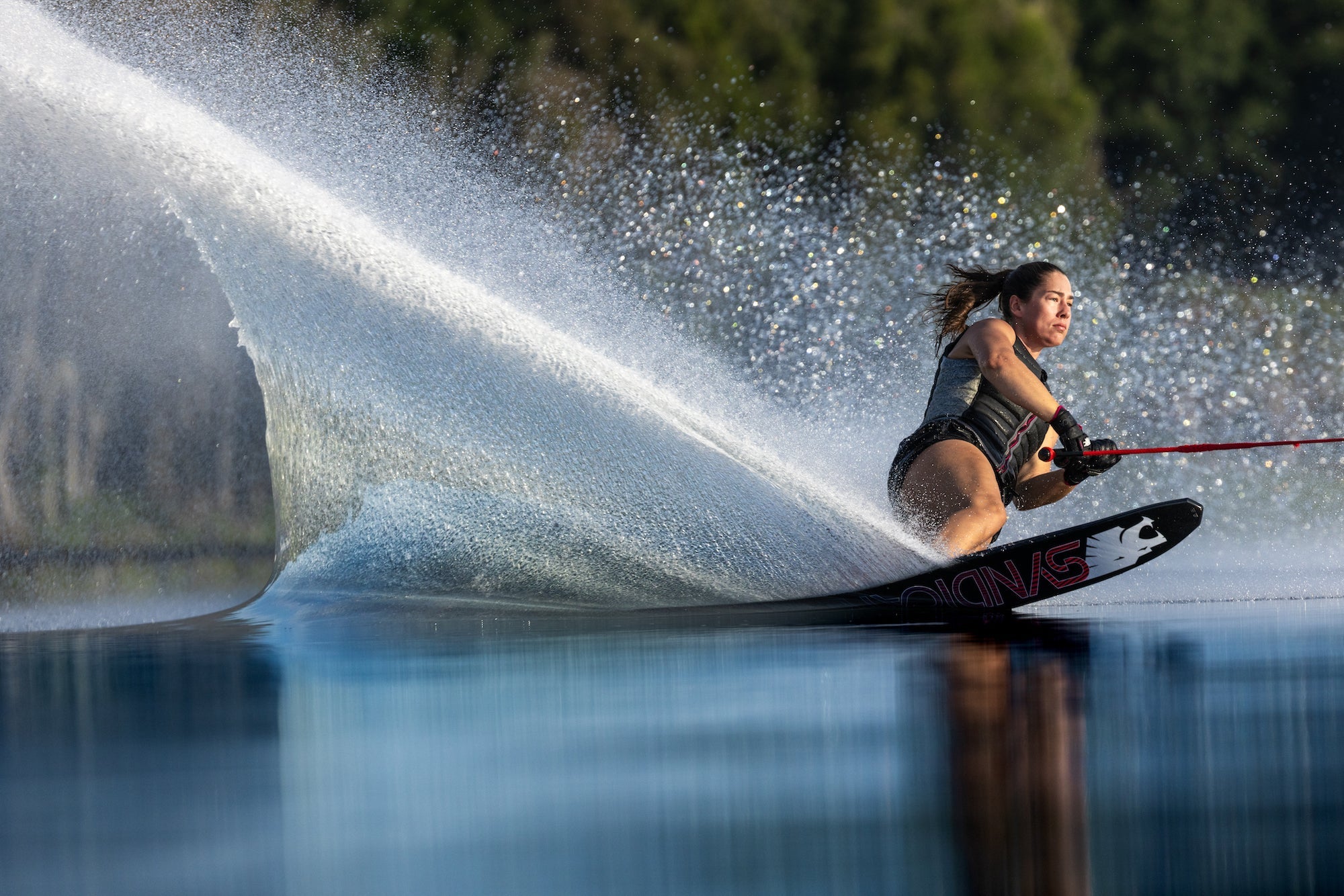 Female Water Skier making a turn and throwing up a dark blue wall of water riding a HO Sports water ski and HO Sports Water Vests.