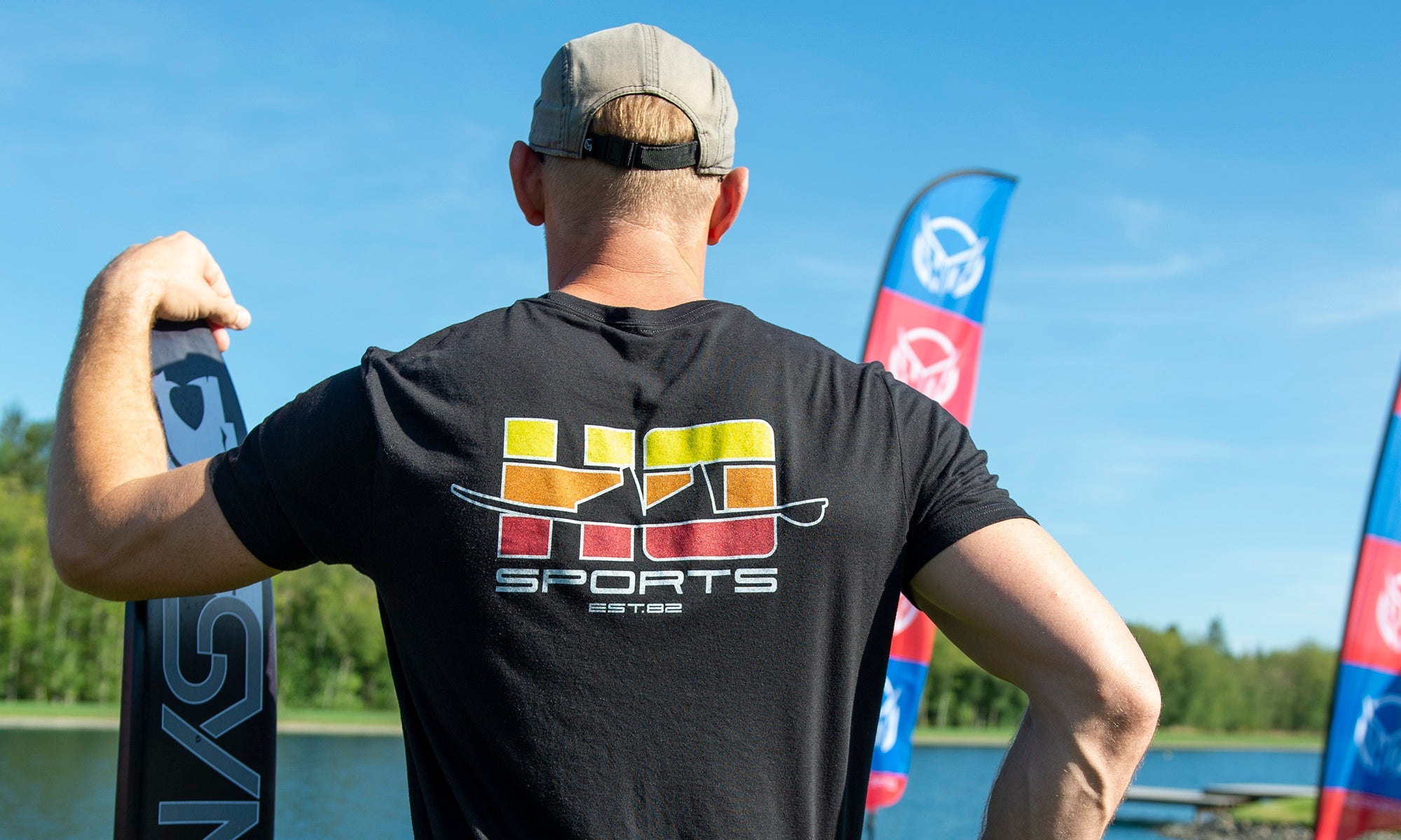 A man looking out over a body of water wearing a black HO Sports T-Shirt with a Yellow, Orange and Red logo. He is holding an HO Sports Syndicate water ski.