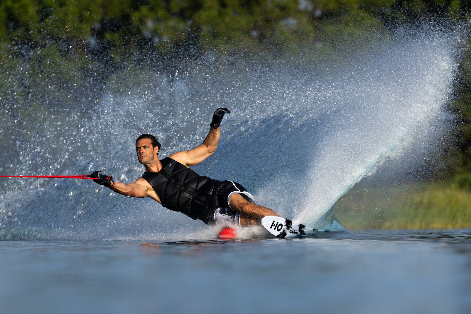 A male waterskier in a black vest and shorts carves through the water, creating a large spray as he leans into the turn.