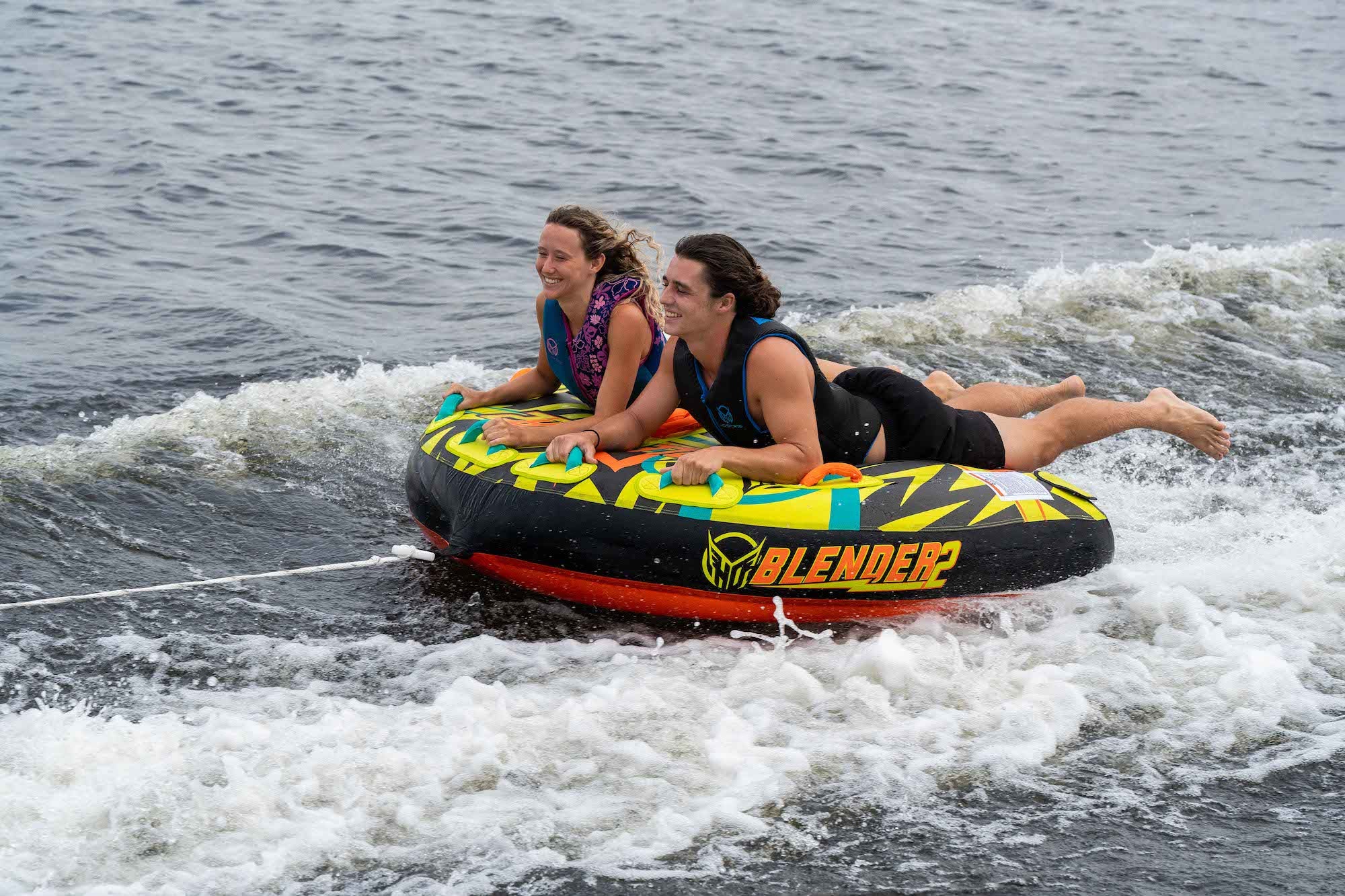Two riders in life vests on a colorful HO Sports Blender 2 towable tube, skimming across the water.