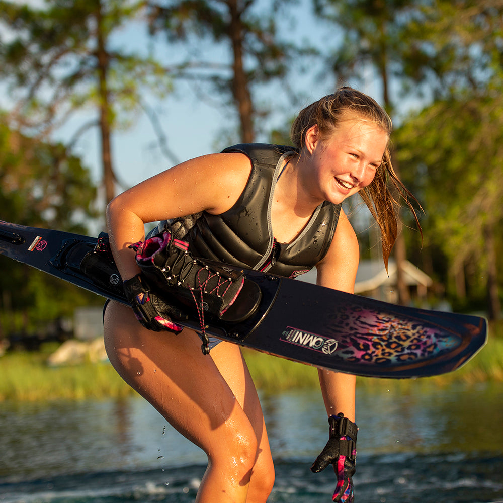 A smiling female waterskier in a black vest and gloves holds a black Omni waterski with pink leopard print graphics.