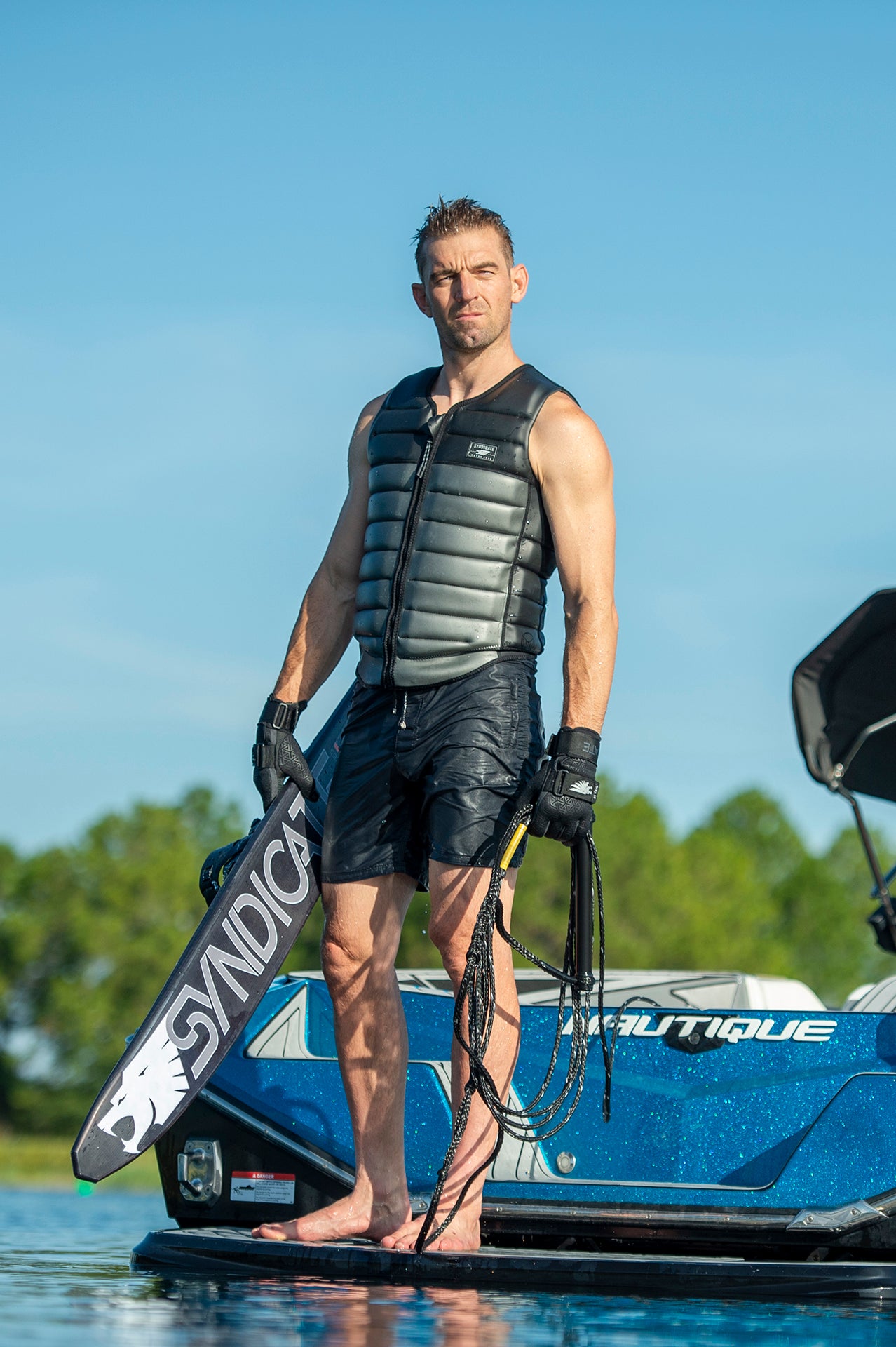 Man in black vest stands on boat platform holding a Syndicate ski and rope handle.