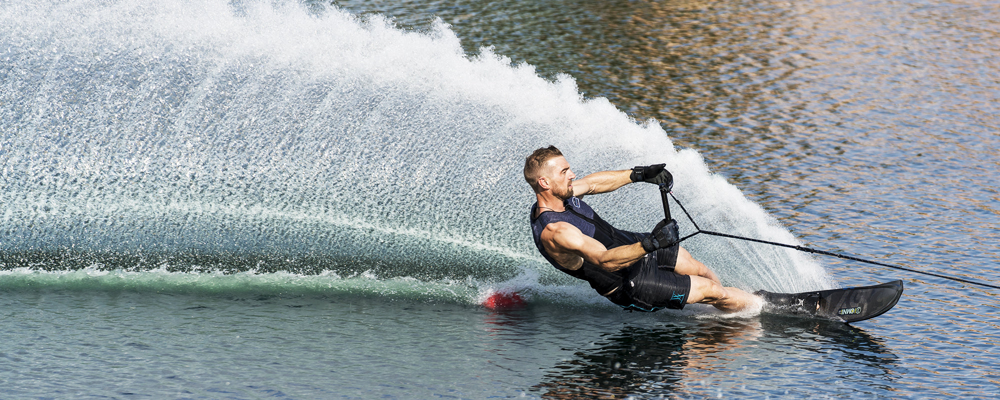 Water skier in a black vest leans into a sharp turn, carving through the water and creating a wide arc of spray.