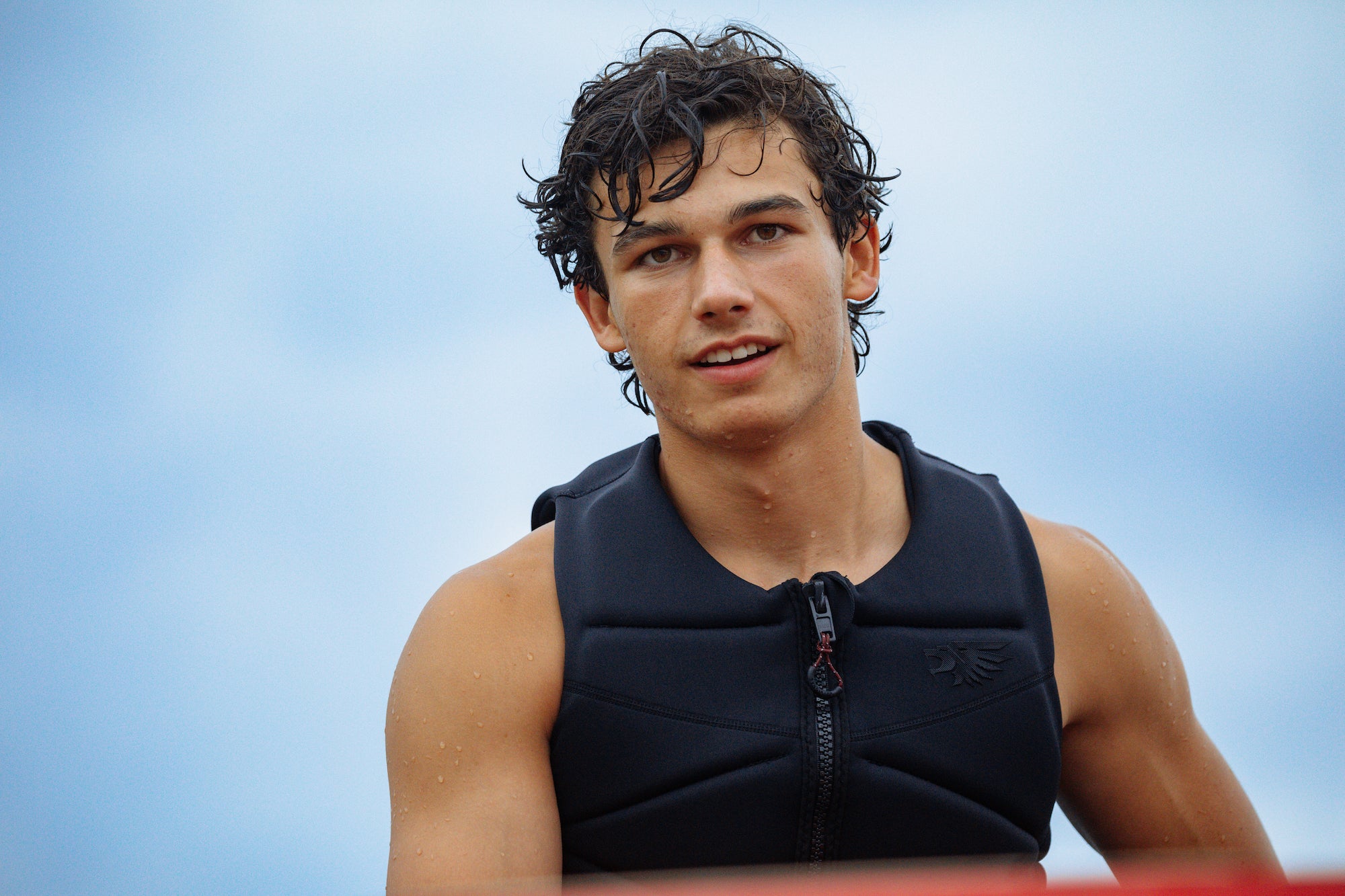Young man in a black vest stands wet and smiling after skiing, blue sky in the background.