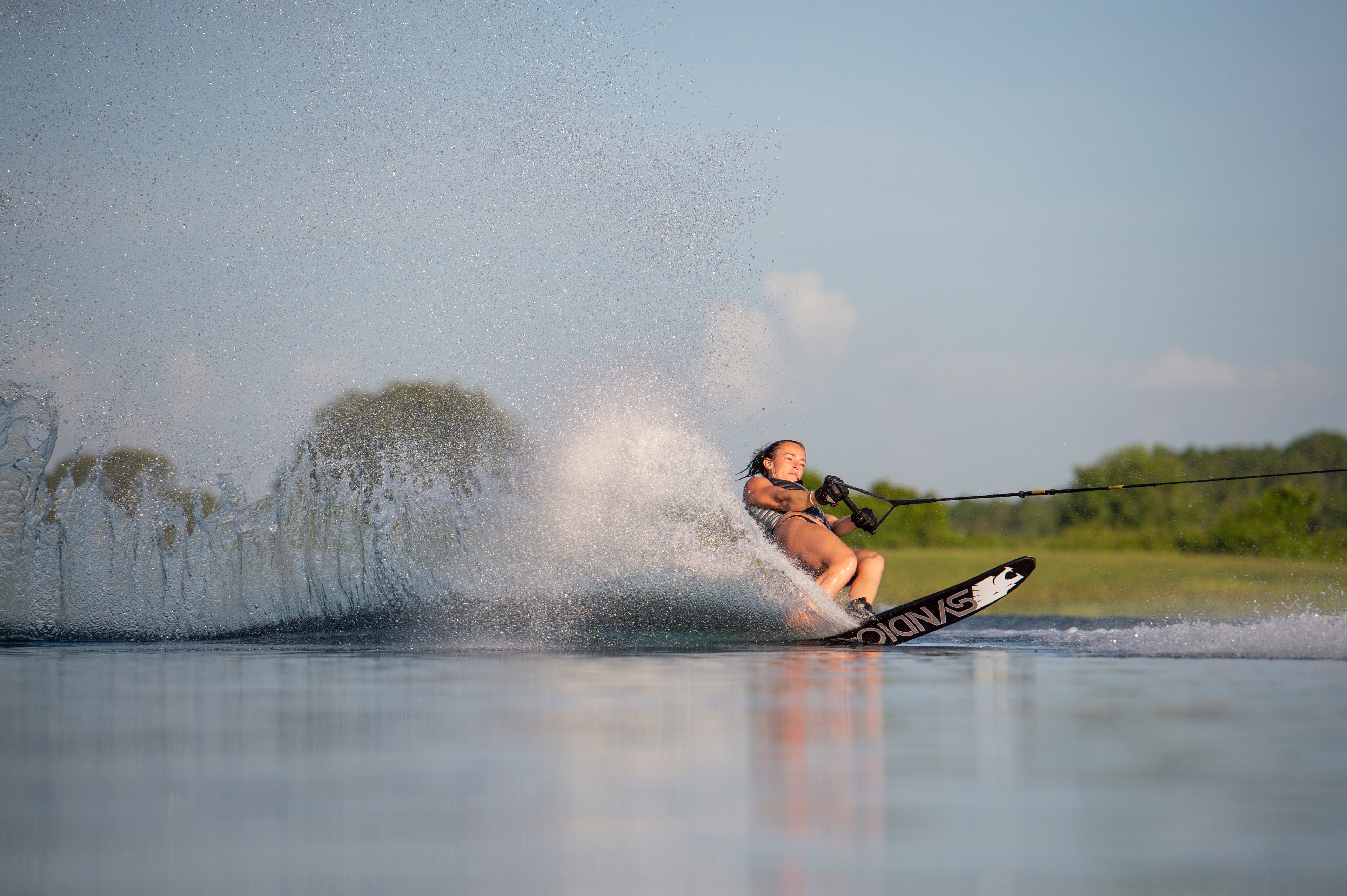 Allie Nicholson cuts across the water on an HO Syndicate ski, creating a high spray against a blue sky.
