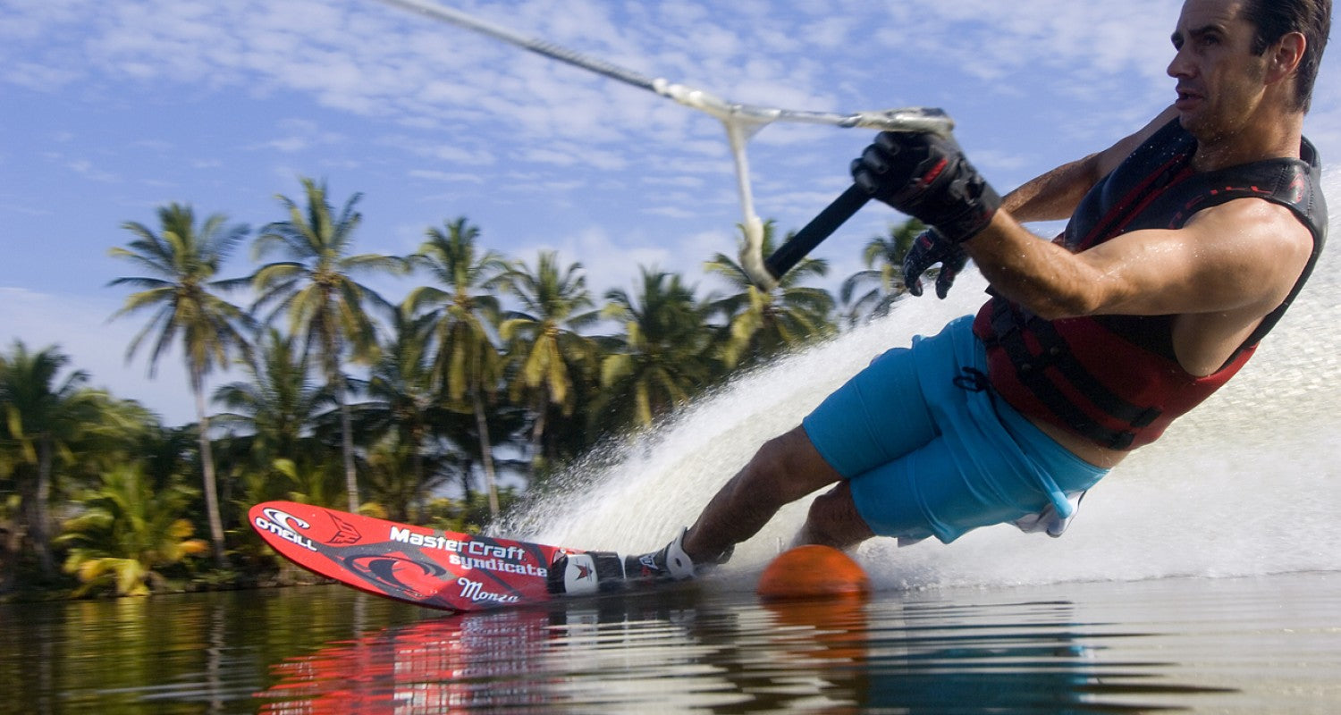 Wade Cox carving through the water on a red slalom ski, with tropical palm trees in the background.