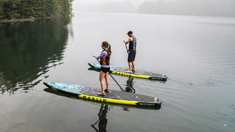 Two paddlers wearing life vests on HO Sports Marlin inflatable paddleboards in a calm, misty lake.